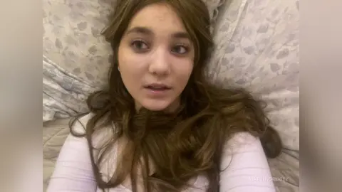 MartineKibbey with long wavy brown hair, wearing a white top while lying on a floral pillow.