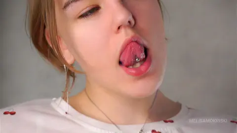 Close-up of a young woman with a tongue piercing wearing a white floral shirt in a soft, natural light setting.