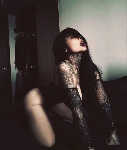 Tattooed woman in black lingerie and fishnet gloves posing moody in a dimly lit room.