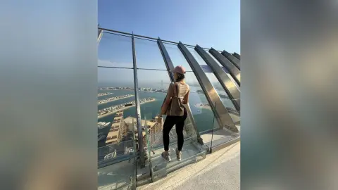 Woman in casual tank top and leggings standing on a high-rise balcony overlooking a coastal city skyline.