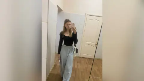Young woman in a black long-sleeve top and grey wide-leg trousers taking a mirror selfie in a minimalist room.