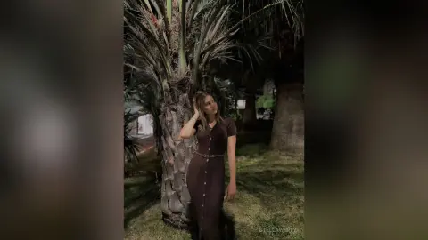 Elegant woman in a brown midi dress posing by a palm tree in a moody night garden setting.