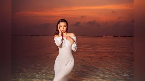 Elie poses in a tight white dress against a sunset ocean background, touching her face with one hand.