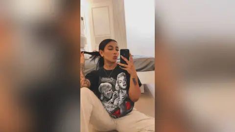 Young woman in a black graphic t-shirt and white pants taking a selfie in a bright bedroom.