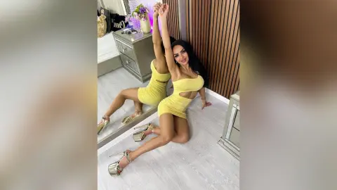 Glamorous woman in a tight yellow mini dress and gold heels posing in a modern bedroom.
