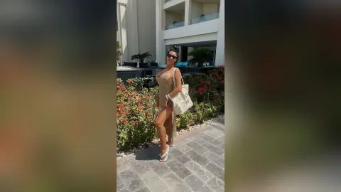 MissyJolie stands outdoors in a tan cut-out bodysuit and sunglasses, posing next to a flower bed near a white building.