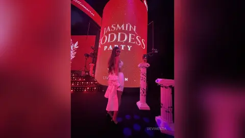 DeviNessa stands in front of a large red backdrop, wearing a white outfit and posing near white decorative pillars.