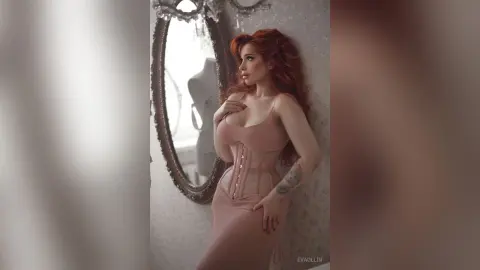 Elegant redhead woman in a blush pink corset dress posing by an antique mirror in a dreamy, vintage setting.