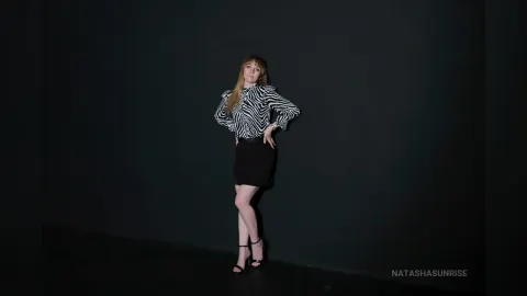 NatashaSunrise — NatashaSunrise poses in a black and white striped shirt and a short black skirt against a dark background.