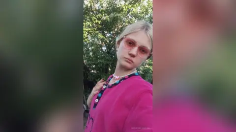 GeorgineAragan wears pink-tinted sunglasses and a bright pink button-up shirt while standing outdoors against a background of green trees.