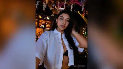 Young woman in a white button-down shirt posing in a dimly lit, moody lounge setting.