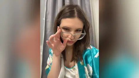 Young woman in a teal satin robe wearing clear glasses in a bright indoor setting with a playful mood.