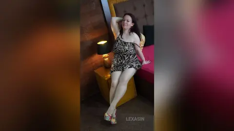 Smiling woman in a leopard print mini dress posing in a cozy bedroom setting.