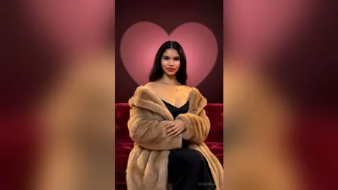 Elegant woman in a black dress and tan faux fur coat posing against a romantic heart backdrop.