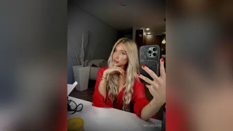 Blonde woman in a red silk blouse taking a mirror selfie in a modern living room with a chic, moody vibe.