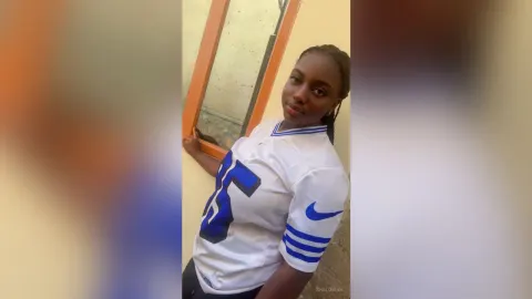 Shaluwah — Shaluwah poses outdoors in a white jersey with blue stripes, leaning against a window frame.