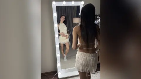Young woman in a white sleeveless mini dress posing in front of a lighted vanity mirror in a bedroom.