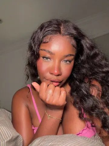 Beautiful Black woman with wavy hair and glowing makeup wearing a pink top in a cozy bedroom setting.