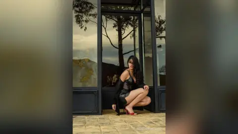 Elegant woman in a black dress posing by a modern glass window against a scenic mountain landscape at dusk.