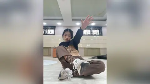 Young woman in a black t-shirt and brown trousers sitting relaxed on a floor in a bright modern room.
