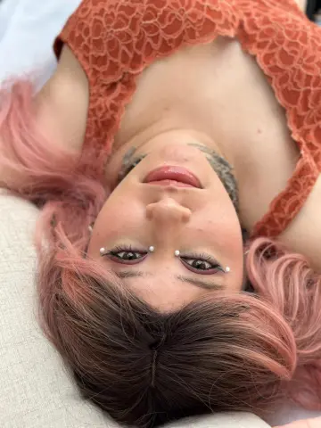 Izzumi lies on a white surface with her pink-tinted hair spread out, wearing an orange lace top.