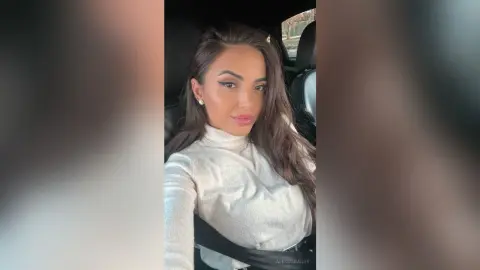 Glamorous woman in a white turtleneck sitting in a car with elegant makeup and long wavy hair.