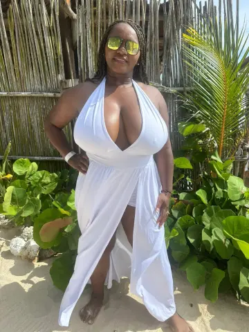 Curvy woman in a white halter maxi dress posing on a sunny tropical beach near palm leaves.