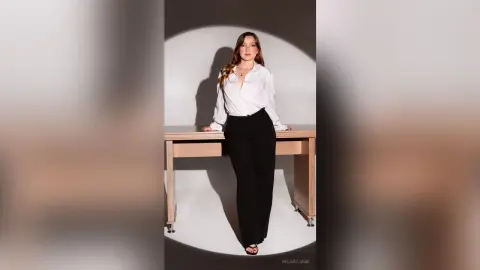 Elegant woman in a white button-down shirt and black trousers posing against a minimalist studio background.