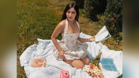 Young woman in a white floral sundress reading a book on a picnic blanket in a sunny green meadow.