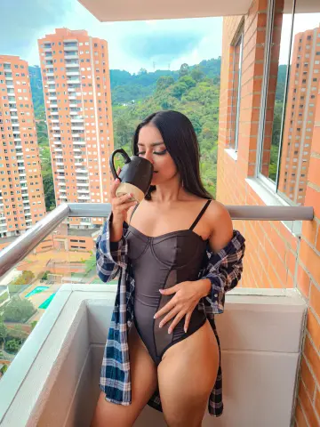 Young woman in a black bodysuit and plaid shirt sipping coffee on a high-rise balcony overlooking city buildings.
