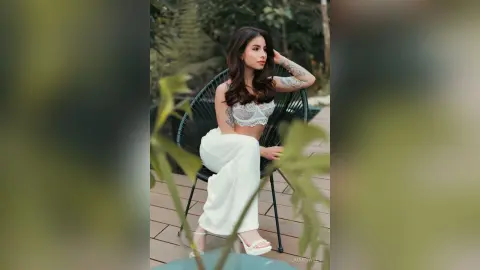 Elegant woman in white lace crop top and flowing trousers relaxing on a patio chair in a lush garden setting.