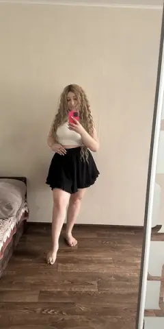 Young woman with wavy blonde hair wearing a white crop top and black skirt posing in a minimalist room.