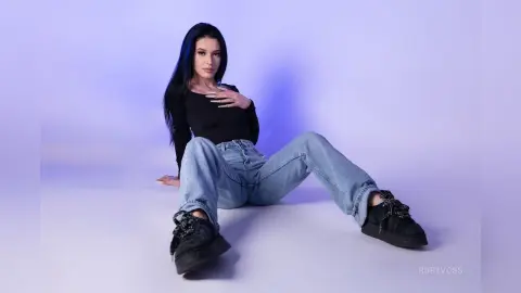 Young woman with long dark hair wearing a black long-sleeve top and light blue jeans sitting against a purple backdrop.