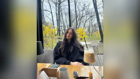 Young woman in a black sweater sitting at a cafe table with iced coffee by a window overlooking autumn trees.