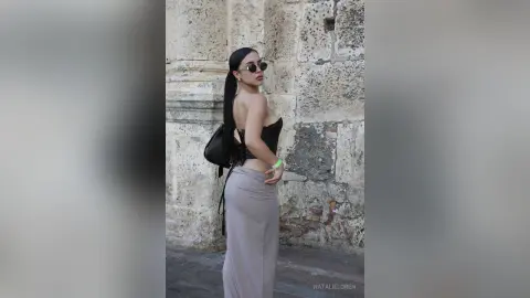Stylish woman in a black corset top and grey maxi skirt posing against a rustic stone wall.