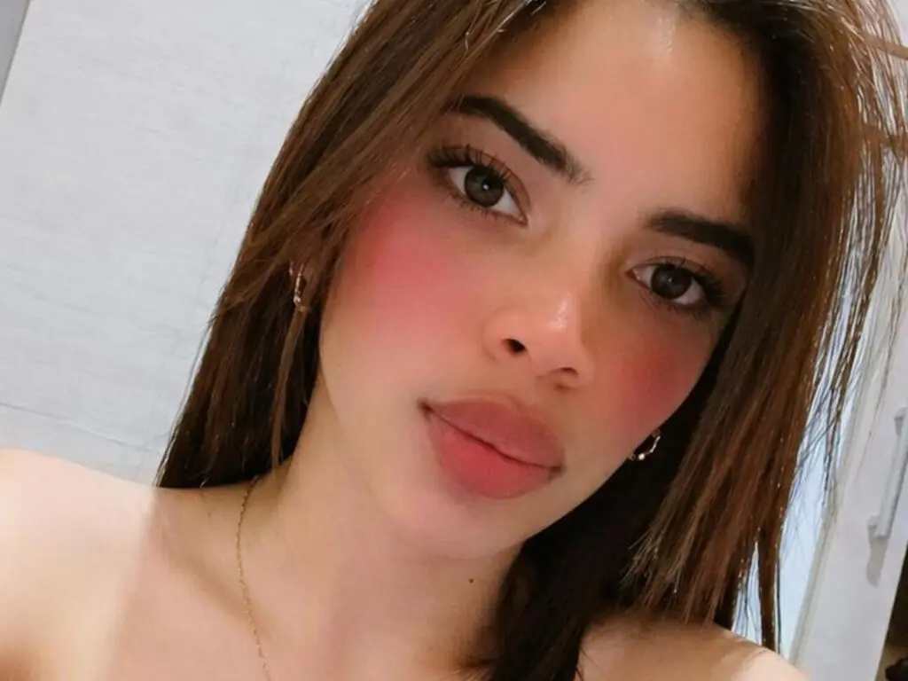 Close-up portrait of a young woman with long brown hair and glowing makeup in a bright indoor setting.