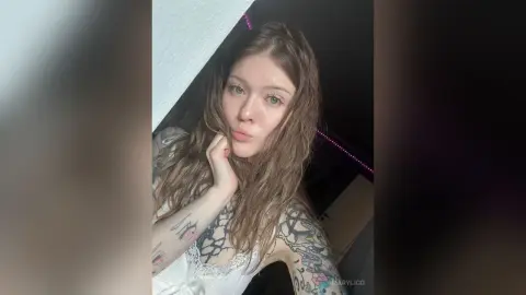MaryLico poses with her hand under her chin, showing her long wavy hair and tattoos on her arms and chest while wearing white lace lingerie.