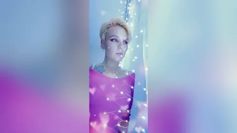AvaLindt poses with blonde hair and heavy eye makeup, wearing a pink top against a background of soft blue light and fairy lights.