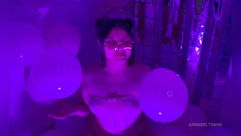 AnnaBeltrann poses in a purple-lit room, wearing a light-colored corset and surrounded by white balloons.
