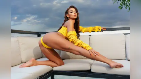 Anastasia poses on a white outdoor sofa wearing a bright yellow lace bikini, kneeling with one leg extended.
