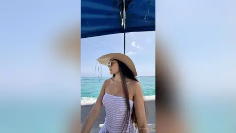 Woman in white strapless top and sun hat relaxing on a boat under a blue canopy by the ocean.