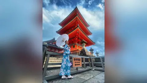 AnaMathers stands outdoors in a blue floral kimono, holding a white paper parasol against a red pagoda.