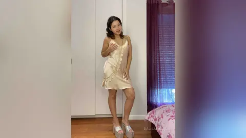 DalilaPearl — DalilaPearl stands in a cream satin slip dress and silver platform heels in a bedroom.