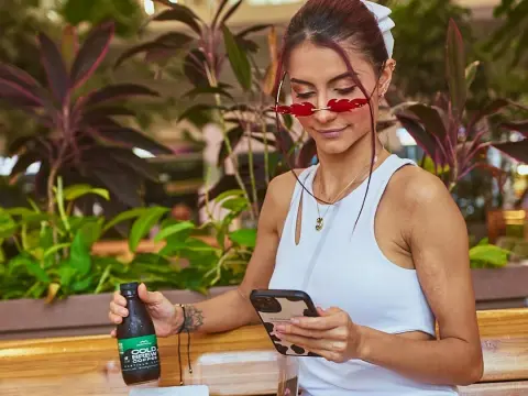 AprilJakovish wearing a white tank top and red sunglasses, looking down at her phone while sitting near green plants.