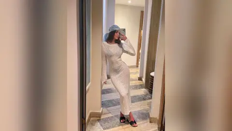 Stylish woman in a white long-sleeve crochet maxi dress posing for a mirror selfie in a modern minimalist hallway.