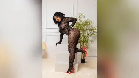 Curvy woman in a black lace bodysuit posing elegantly in a bright, minimalist studio setting.