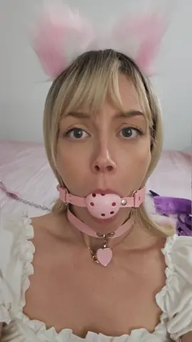 Blonde woman in white ruffled top wearing pink bunny ears and a heart choker in a soft, dreamy bedroom setting.