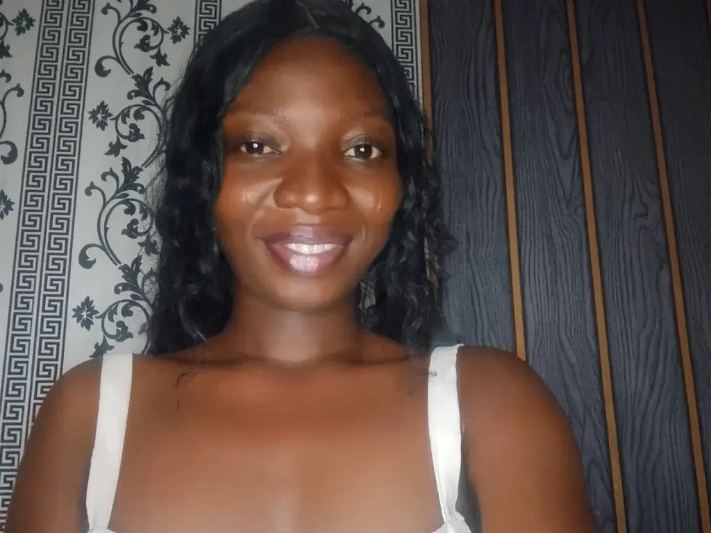 Smiling Black woman in a white tank top posing against patterned wallpaper in a cozy indoor setting.