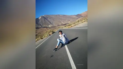 Young man in a light blue shirt and jeans sitting on a desert road with mountains under a clear blue sky.