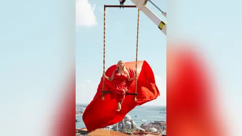Elegant woman in a flowing red dress sitting on a high swing against a bright blue sky background.